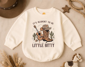 May include: A cream-colored long-sleeve sweatshirt featuring a graphic print of a brown cowboy hat, a brown cowboy boot, an acoustic guitar, and two cacti. The text "IT'S ALRIGHT TO BE LITTLE BITTY" is printed in brown above and below the graphic.