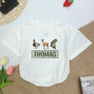 May include: White short-sleeved t-shirt with a green and white plaid border around the text "THOMAS". The shirt features a duck, deer, and fish graphic.