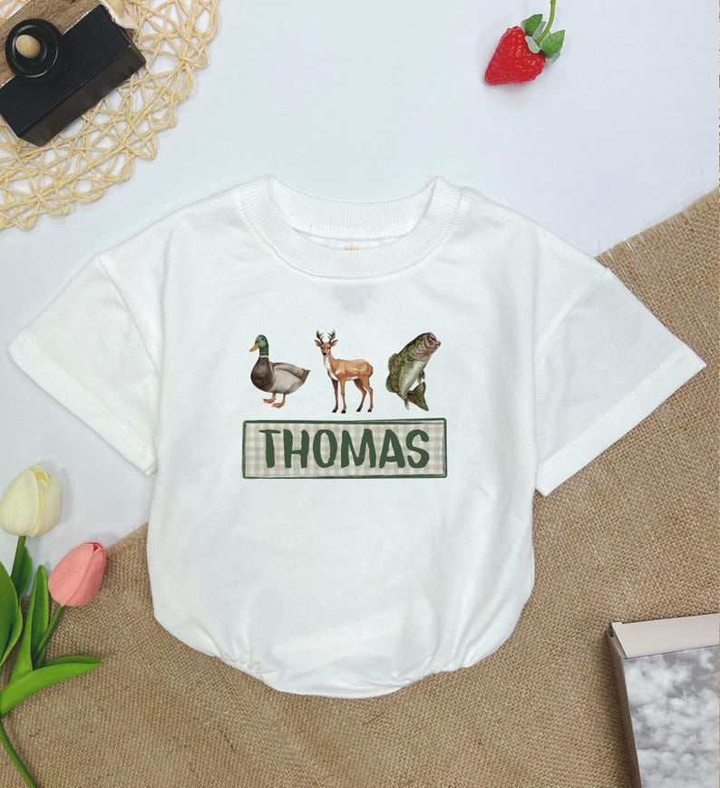 May include: White short-sleeved t-shirt with a green and white plaid border around the text "THOMAS". The shirt features a duck, deer, and fish graphic.