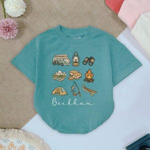 May include: Teal short-sleeved baby bodysuit with a camping-themed graphic. The design includes a camper van, lantern, binoculars, mountains, s'mores, campfire, tent, fishing rod, and canoe. The name "Beckham" is written in cursive below the illustrations.