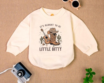 May include: A cream colored long-sleeved onesie with a graphic of a cowboy boot, a guitar, and cacti. The text on the onesie reads "It's alright to be little bitty".
