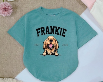 May include: A teal blue short-sleeved t-shirt with a black graphic of a golden retriever dog peeking over a banner that reads "FRANKIE EST 2025".