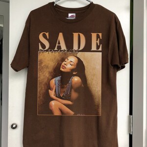 May include: Brown t-shirt with a graphic print of Sade, the singer, with the text "SADE" and "Love Deluxe" in a brown and orange font.