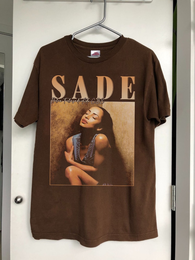 May include: Brown t-shirt with a graphic print of Sade, the singer, with the text "SADE" and "Love Deluxe" in a brown and orange font.