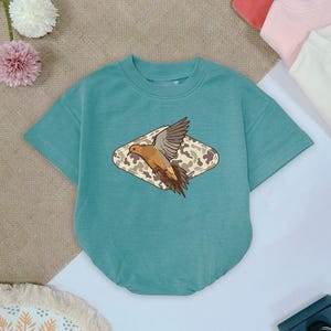 May include: A teal short-sleeved baby bodysuit with a crew neck. The bodysuit features a graphic of a bird with outstretched wings inside a diamond-shaped camouflage design. Other pastel-colored baby clothes are visible in the background.