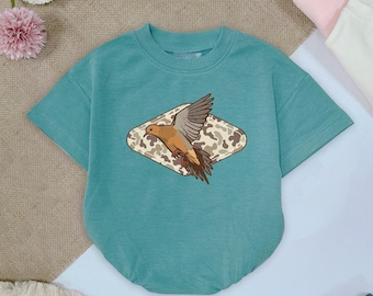 May include: A teal short-sleeved baby bodysuit with a crew neck. The bodysuit features a graphic of a bird with outstretched wings inside a diamond-shaped camouflage design. Other pastel-colored baby clothes are visible in the background.