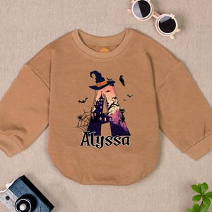 May include: A brown long-sleeved sweatshirt with a Halloween-themed graphic of a witch's hat, a castle, and bats. The text "Alyssa" is printed below the graphic.