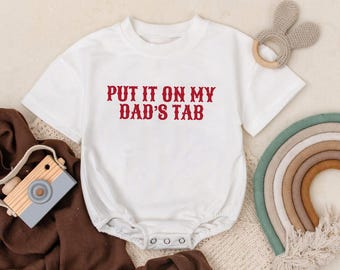 May include: White baby onesie with the text "PUT IT ON MY DAD'S TAB" in red. The onesie has short sleeves and snaps at the bottom. A wooden toy camera, a rainbow, and a bunny-shaped teether are also in the image.