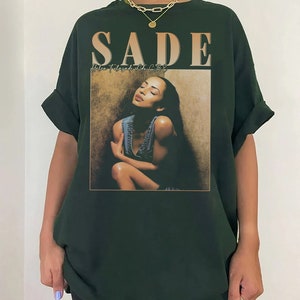 May include: A dark green t-shirt with a graphic print of Sade, a singer, with the text "SADE" and "Helen Folasade Adu CBE" printed on the front.