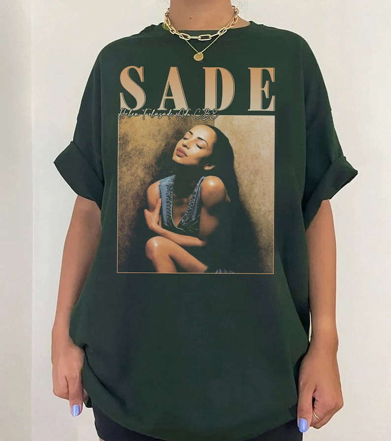 May include: A dark green t-shirt with a graphic print of Sade, a singer, with the text "SADE" and "Helen Folasade Adu CBE" printed on the front.