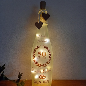 Könnte beinhalten: Eine durchsichtige Glasflasche mit einer goldenen Aufschrift, die "Goldene Hochzeit 50 Jahre Rosemarie & Armin 06.11.1972" lautet. Die Flasche ist mit zwei kleinen Holzherzen und einer Lichterkette verziert.