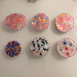 May include: Six decorative resin phone grips in various shapes, including round and hexagonal. The grips feature glitter and decorative elements in pink, blue, gold, and silver. Accents include stars and butterflies, creating a colourful, eye-catching design.