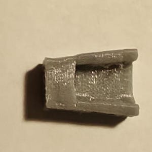May include: A small, gray, rectangular object with a hollowed-out center. The object has a textured surface and appears to be made of a solid material. The object is casting a shadow on a light-colored surface.