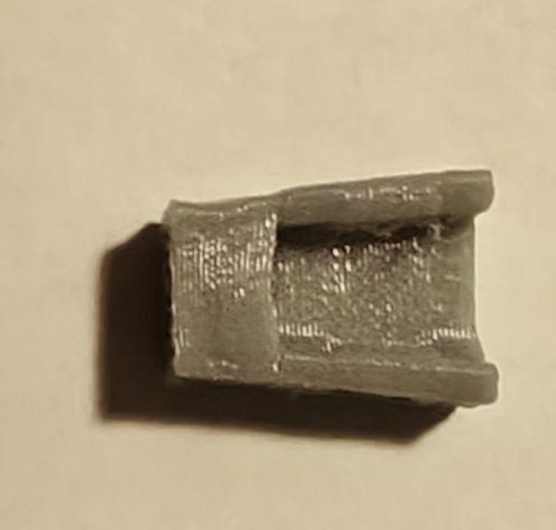 May include: A small, gray, rectangular object with a hollowed-out center. The object has a textured surface and appears to be made of a solid material. The object is casting a shadow on a light-colored surface.