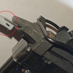 May include: Close-up of a nail gun mechanism. The image highlights a small, white plastic component with a rectangular shape, positioned at the end of the gun's metal structure. The nail gun is black and gray.