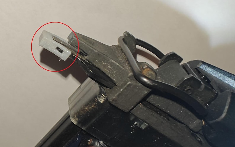 May include: Close-up of a nail gun mechanism. The image highlights a small, white plastic component with a rectangular shape, positioned at the end of the gun's metal structure. The nail gun is black and gray.