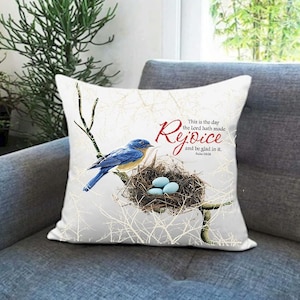 May include: White cushion with a bluebird perched on a branch with a nest of blue eggs. The cushion has the text "This is the day the Lord hath made Rejoice and be glad in it. Psalm 118:24"