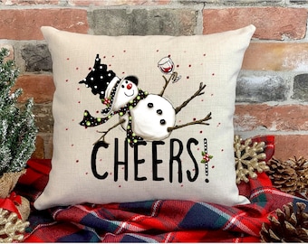Könnte beinhalten: Ein weißer Schneemann mit einem schwarz-weißen Hut und einem grünen Schal hält ein Glas Rotwein. Der Schneemann sitzt auf einem beigefarbenen Kissen mit dem Wort "CHEERS!" in Schwarz gedruckt.