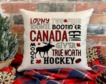 May include: A white throw pillow with a red, black, and white design. The pillow features the words "Loony Two Four & Toonie Bootin'er Canada Moose Give'r True North Hockey" with a moose silhouette and a maple leaf.