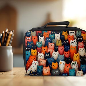 May include: Black lunch bag with a vibrant design of cartoon cats in diverse colours. The bag has a black handle and zip. A pencil pot with pencils is to the left of the bag.