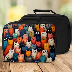 May include: Black lunch bag with a colourful cat design. The front of the bag features a vibrant illustration of numerous cats in various colours, including orange, blue, and white. The bag has a black handle and a solid black back.