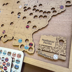 May include: Wooden map of the United States with cutouts for states and various stickers. Includes a "National Parks & Ballparks" sign and stickers with baseball and park logos. The map is framed in light wood.