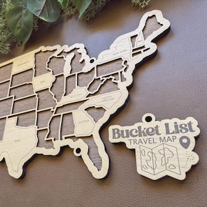 May include: Wooden United States map with state outlines, plus Alaska and Hawaii, and a separate "Bucket List Travel Map" tag. The map is made of light-colored wood with a dark wood backing. The states are labeled with their names.