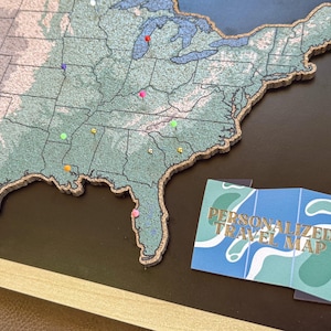 May include: A framed map of the United States with colorful pins marking locations. The map is a textured green and blue with a cork border. A small card with the text "PERSONALIZED TRAVEL MAP" is in the lower right.