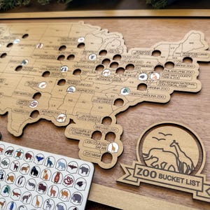 May include: Wooden map of the United States with zoo locations marked. Includes a sticker sheet with animal icons and a wooden sign that says "ZOO BUCKET LIST". The map is framed in a light wood.
