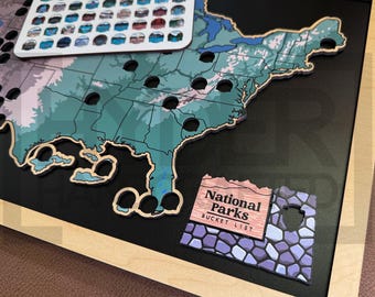 May include: A wooden map of the United States with black circles representing national parks. The map is framed in a light wood frame. A separate wooden sign reads "National Parks Bucket List" with a stone-like pattern.