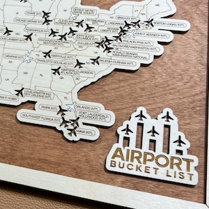 May include: A wooden map of the United States with the names of major airports printed on it. The map is surrounded by a light brown frame. The text "AIRPORT BUCKET LIST" is printed on a separate wooden piece below the map.