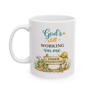May include: White ceramic mug with the text "God's still working on me" in teal script. The mug features construction-themed illustrations, including a hard hat, tools, and a sign that reads "Under Construction."