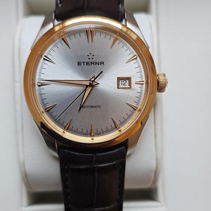 May include: A silver-faced wristwatch with gold accents and a brown leather band. The watch face features the word "ETERNA" and the text "AUTOMATIC". The watch has a date window at the 3 o'clock position.