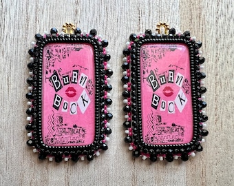 May include: A pair of black and white beaded rectangular pendants with a pink background and the text "Burn Book" in white letters. The pendants have a gold loop at the top.
