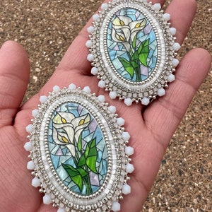 May include: Two oval brooches with a beaded design. Each brooch showcases a stained-glass style image of white calla lilies with green leaves on a blue and white backdrop. They are framed with silver beads and white, faceted beads.