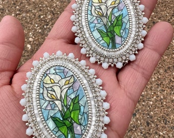 Op de afbeelding: Twee ovale, geborduurde broches. Elke broche heeft een glas-in-lood ontwerp van witte calla lelies met groene bladeren tegen een blauw-witte achtergrond. De broches zijn omlijst met zilveren kralen en witte, facetgeslepen kralen.