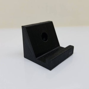 May include: A black plastic phone stand with a hole in the back for mounting. The stand has a small lip on the front to hold a phone in place.
