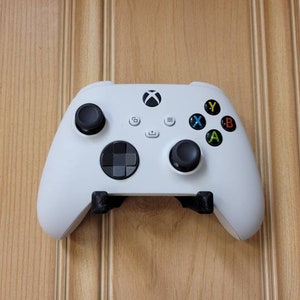 May include: A white Xbox Series X controller with black buttons and a black directional pad. The controller is mounted on a wall.