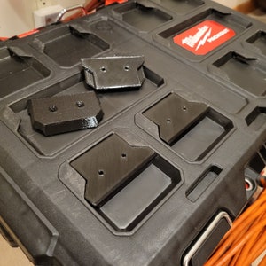 May include: Four black plastic tool holders with screw holes, sitting on a black toolbox with the Milwaukee logo and the word "PACKOUT" printed on it.