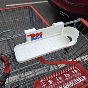 May include: A white plastic "The Dog Dock" shopping cart accessory. It features a tray with grooves and a cup holder. The product is designed to attach to a shopping cart. The text "THE DOG DOCK" is printed on the product.