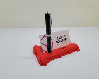 May include: Red 3D printed desk organizer shaped like a car engine block with a black pen and a business card that reads "Zandelay Industries 3D Printing and Design Solutions" in a white and pink color scheme.