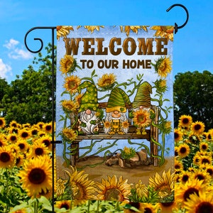 Puede incluir: Una bandera de jardín de bienvenida con tres gnomos sentados en un banco. Los gnomos llevan sombreros verdes y tienen barbas blancas. La bandera está rodeada de girasoles y tiene el texto "Bienvenido a nuestro hogar".