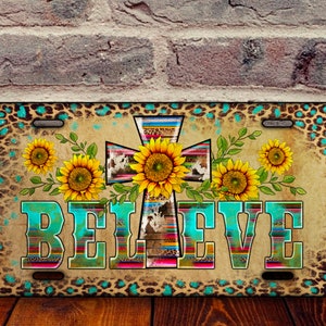 May include: A turquoise and brown license plate with a rustic background featuring a cross with a southwestern pattern and sunflowers. The word "BELIEVE" is written in a turquoise and brown font.