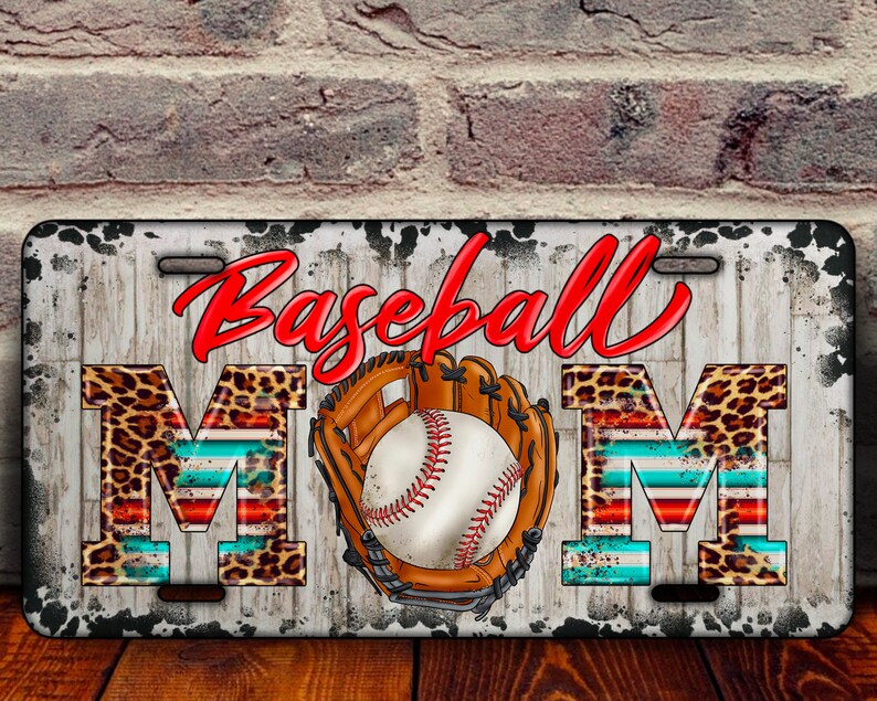 K&ouml;nnte beinhalten: Ein wei&szlig;es Nummernschild aus Metall mit einem abgenutzten Look. Die Worte "Baseball Mom" sind in roten und schwarzen Buchstaben mit Leopardenmuster und rot-wei&szlig;-blauen Streifen. Ein Baseball und ein Handschuh befinden sich in der Mitte des Nummernschilds.