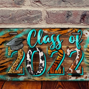 May include: A wooden license plate with a turquoise border and a graduation cap. The numbers 2022 are in the centre with a leopard print, cow print, and tiger print design. The text "Class of" is written in cursive above the numbers.