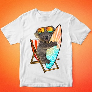 May include: White t-shirt with a graphic of a koala wearing sunglasses and holding a surfboard. The koala is sitting in a beach chair. The graphic is in a cartoon style.
