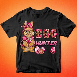 May include: Black t-shirt featuring a cartoon bunny wearing glasses and a pink floral patterned outfit. The text "EGG HUNTER" is in pink and brown camouflage print. Three decorated Easter eggs are also shown.