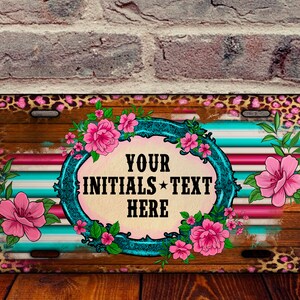 May include: Rectangular license plate with a wood grain background, pink and turquoise stripes, and a leopard print border. The center features a floral design with the text "YOUR INITIALS TEXT HERE". Pink flowers and green leaves are also present.