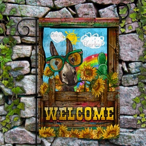 Op de afbeelding: Een welkomst-tuin vlag met een cartoon ezel die een zonnebril draagt en een regenboog achter zich heeft. De ezel is omringd door zonnebloemen en een cactus. De vlag heeft een houten frame en het woord "WELCOME" in gouden letters.