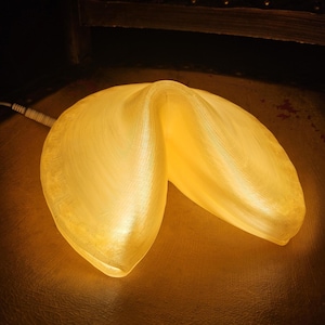 Op de afbeelding: Een lamp in de vorm van een gelukskoekje die een warm, zacht geel licht uitstraalt. Het doorschijnende, getextureerde oppervlak van de lamp bootst het uiterlijk van een gebakken gelukskoekje na. Een wit netsnoer steekt uit de linkerkant.